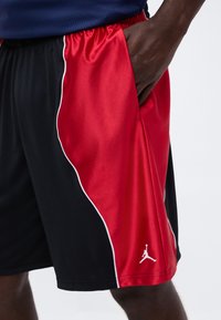 Primer plano de una persona que lleva pantalones cortos de baloncesto negros y rojos con ribete blanco y un logo blanco de Jumpman cerca del dobladillo, mano en el bolsillo derecho.
