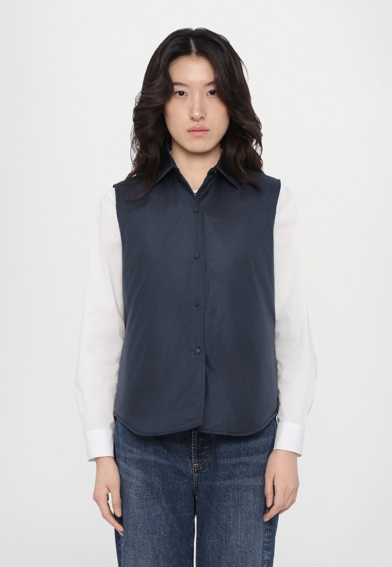 Navy mouwloze vest met een kraag, knopen aan de voorkant en een gladde textuur, gedragen over een wit shirt met lange mouwen en gecombineerd met denim jeans.