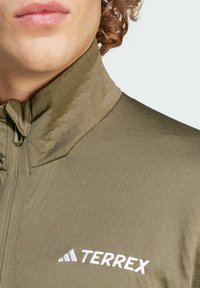 Giacca verde oliva con colletto alto in un tessuto testurizzato. Presenta una zip anteriore e il logo "TERREX" ricamato in bianco sul petto.