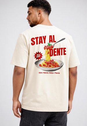 Mann trägt ein beiges T-Shirt mit einer Gabel, die Spaghetti anhebt, und den Text "STAY AL DENTE ALL DAY Eat pasta move fasta" auf dem Rücken.