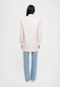 Mulher com cabelo longo e escuro, vestindo uma camisa de manga longa listada e jeans azuis com pequenos padrões florais rosa, de pé de costas.