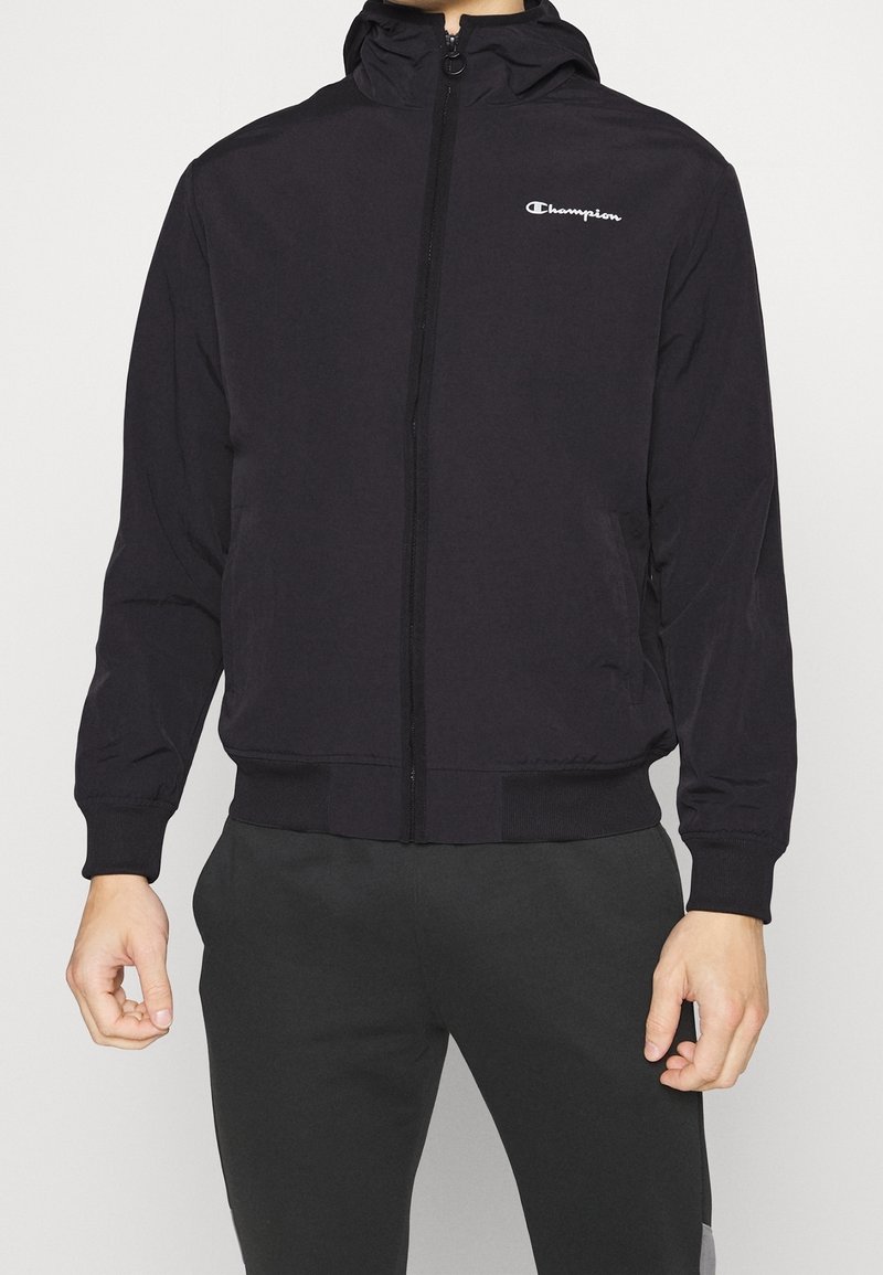 Black zip-up jacket with hood, featuring a logo on the chest, elastic cuffs, and a smooth fabric texture. Accented by a sporty design.