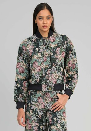 Femme portant un ensemble assorti de veste bomber et pantalon à imprimé floral avec des poignets et une taille côtelés noirs, debout devant un fond gris uni.
