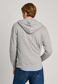 Sudadera gris con capucha ajustable y puños acanalados, superpuesta a una camisa de botones azul claro con rayas blancas, combinada con jeans azul oscuro.