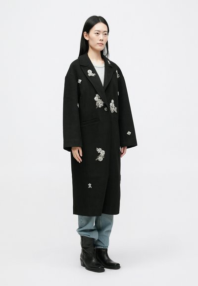Črn oversize plašč z cvetličnim vezenjem, odprt spredaj, z velikimi klapnicami in stranskimi žepi, v kombinaciji z modrimi kavbojkami in črnimi gležnjarji.