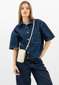 Camicia di jeans a maniche corte, colore blu scuro, con tasche laterali e chiusura a bottoni. Abbinata a una borsa a tracolla color crema.