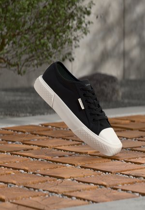 Schwarzer Sneaker mit niedrigem Schaft, weißer Sohle und Zehenkappe schwebt über Holzkacheln im Freien nahe einem grünen Busch und einer Betonwand.