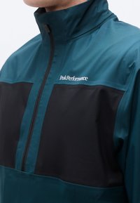 Άνδρας που φοράει τζάκετ με φερμουάρ σε τιρκουάζ και μαύρο, με ψηλό γιακά και λογότυπο "Peak Performance" στο στήθος.