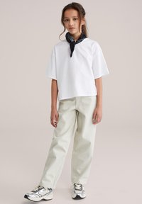 Ung pige iført en hvid oversized t-shirt, lysebeige bukser, sølvfarvede sneakers og et sort mønstret halstørklæde, stående foran en ensfarvet baggrund.