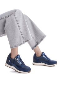 Zapatillas deportivas azules con materiales de ante y sintéticos, que cuentan con un cierre metálico, cordones planos y una entresuela blanca, combinadas con pantalones grises.