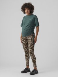 Umstandst-Shirt in Dunkelgrün mit kurzen Ärmeln und seitlicher Raffung, kombiniert mit Leggings im Leopardenmuster. Schwarze Plateauschuhe.