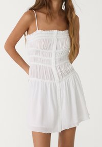 Vestido blanco sin mangas con una textura fruncida, que presenta un botón en la parte delantera y un dobladillo acampanado. Tela suave y ligera.