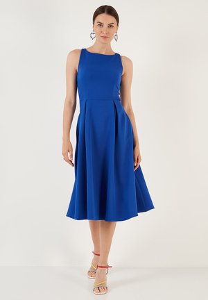 Žena v bezrukávových kráľovskomodrých šatách po kolená s plisovaním, v kombinácii s viacfarebnými sandálmi na vysokom opätku s prackami a kruhovými náušnicami.