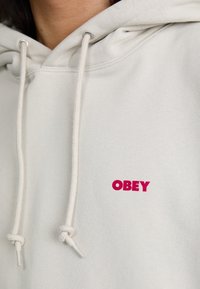 Light gray hoodie with a drawstring and a small red "OBEY" logo on the chest. Features a soft texture and simple design.
