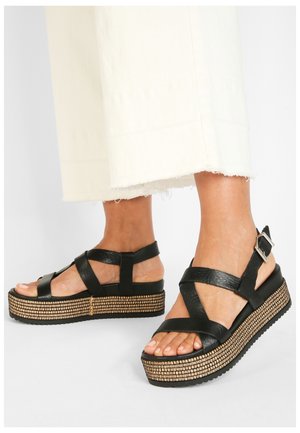 Sandales compensées en cuir noir avec design à lanières croisées, plateforme texturée et appliques argentées sur la bride de cheville. Modèle avec bout ouvert.