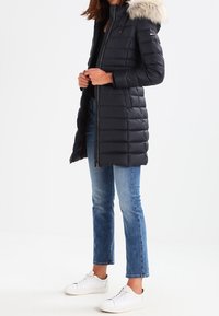 Black puffer coat with a fur-lined hood, zip closure, and quilted design. Worn with light blue jeans and white sneakers.