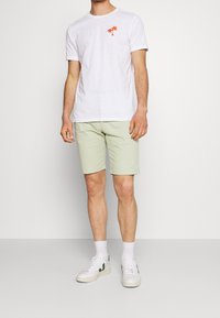 T-shirt bianca a maniche corte con grafica di palma arancione, abbinata a pantaloni corti verde chiaro e sneaker bianche con dettagli verdi.
