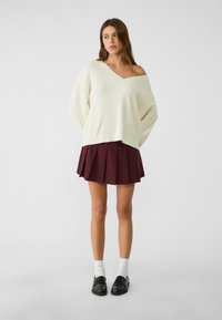 Krémový oversized pletený svetr s výstřihem do V, zkombinovaný s burgunďskou plisé mini sukni. Černé mokasíny a bílé ponožky ke kotníkům dotvářejí look.