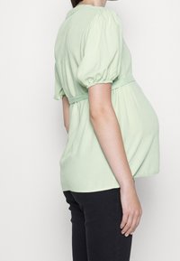 Haut de maternité vert clair avec des manches bouffantes, taille cintrée et tissu fluide. Associé à un pantalon noir, mettant en avant le confort et l'ajustement.