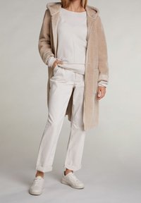 Kvinde iført en beige hættetrøje af fleece, hvid v-hals sweater, hvide bukser med opbrettede sømme og hvide sneakers, der står mod en lys baggrund.