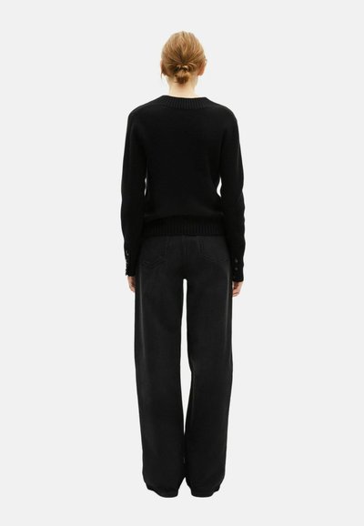 Zwart gebreid sweater met een geribde halslijn en lange mouwen, gecombineerd met wijde zwarte broek. Zichtbare knoopdetails bij de manchetten.