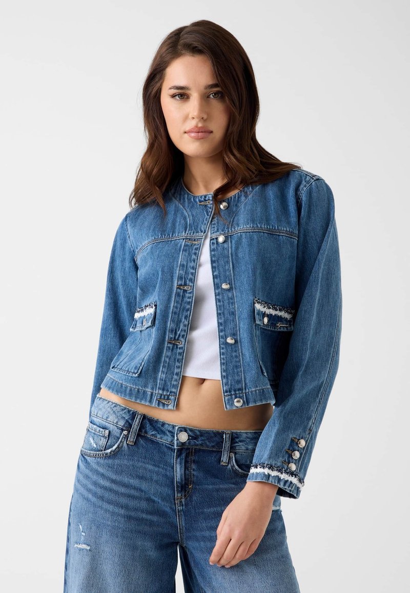 Femme portant une veste courte en jean bleu par-dessus un crop top blanc, associée à un jean bleu, debout devant un fond uni.