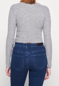 Žena v šedém pleteném topu s dlouhým rukávem a tmavě modrých skinny džínech s vysokým pasem, zobrazená zezadu na jednobarevném pozadí.