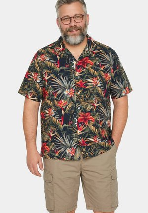 Mann mit grauem Bart und Brille trägt ein schwarzes kurzärmeliges Hemd mit tropischem Muster und beige Cargoshorts, steht mit einer Hand in der Tasche.