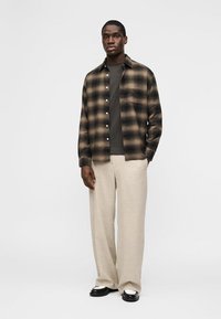 Homme debout portant une chemise à carreaux marron sur un t-shirt foncé, un pantalon beige à jambes larges et des chaussures noires et blanches, sur un fond uni.