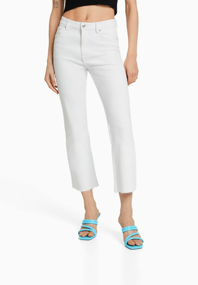 Jean en denim blanc au design raccourci et jambes droites, ourlet effiloché, associé à des sandales à lanières bleues.