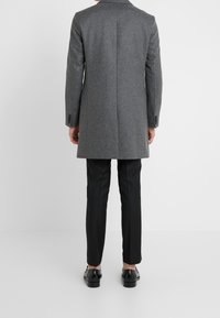 Grijs wollen middenlang overjas met een enkele achterkantopening, gecombineerd met zwarte broek en glanzende zwarte schoenen, gezien van achteren.