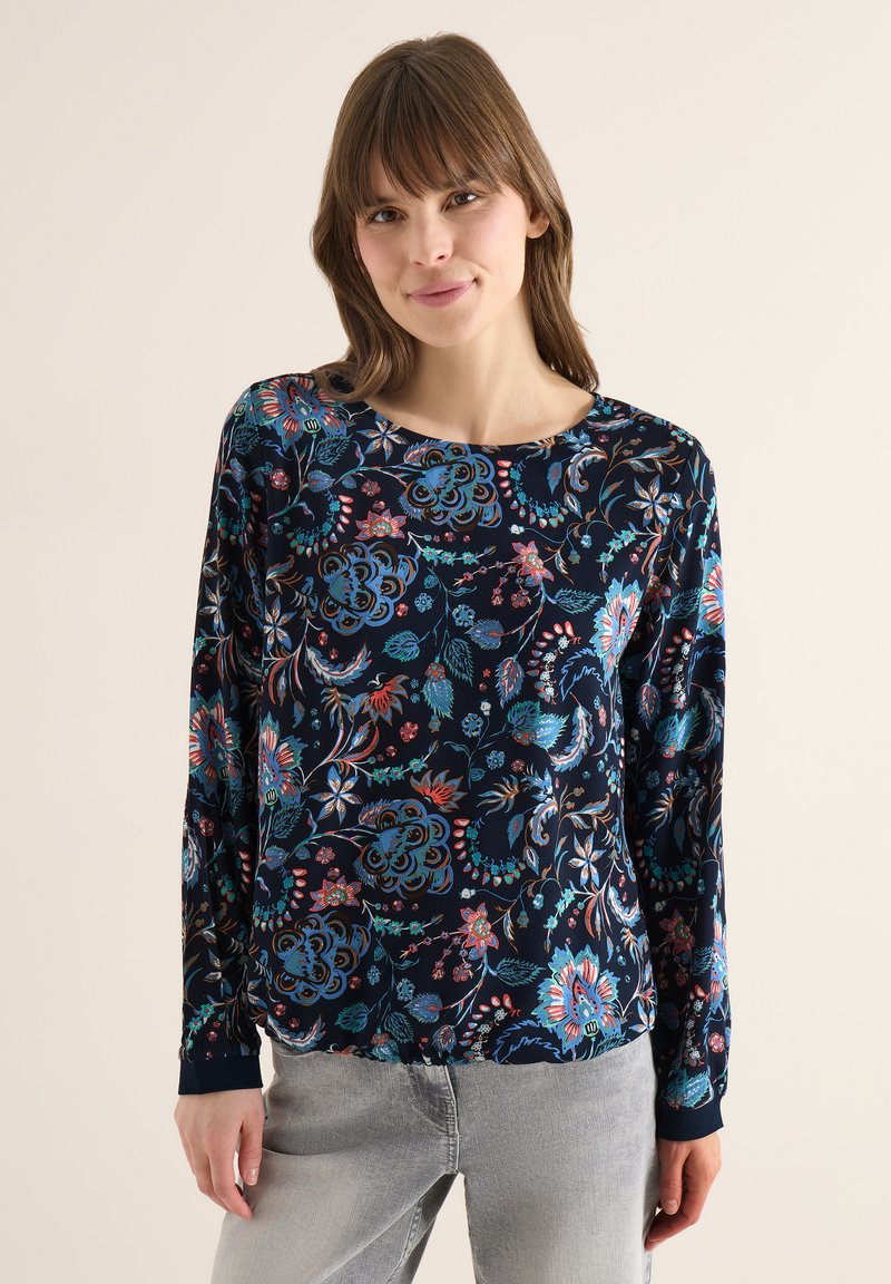 Femme portant un chemisier bleu foncé à manches longues avec des motifs floraux colorés et un jean gris clair, debout devant un fond beige uni.