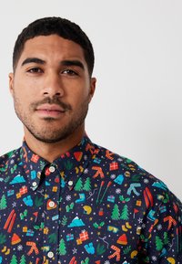 Camicia a maniche corte con bottoni, caratterizzata da uno sfondo blu scuro e motivi colorati a tema invernale, inclusi alberi, regali e fiocchi di neve.