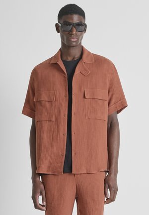 Homme portant une chemise à manches courtes texturée de couleur rouille et un pantalon assorti, un sous-vêtement noir et de grandes lunettes de soleil noires, debout devant un fond gris.