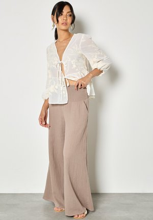 Vrouw met donker haar in een paardenstaart, gekleed in een doorschijnende witte blouse met strik aan de voorkant en bloemenborduurwerk, en beige broek met wijde pijpen, staand tegen een witte muur.