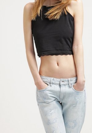 Jeune personne les mains dans les poches d'un jean clair délavé usé, portant un crop top noir sans manches avec une bordure en dentelle sur un fond uni.