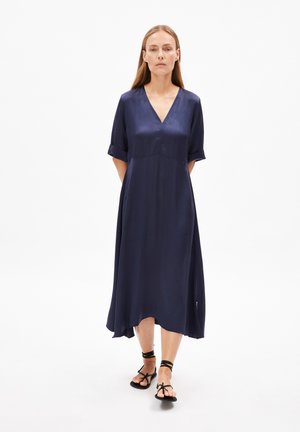 Mörkblå midi-klänning i silkesliknande material, med V-hals, rullade ärmar och en svävande nederkant. Bärs med svarta sandaler.