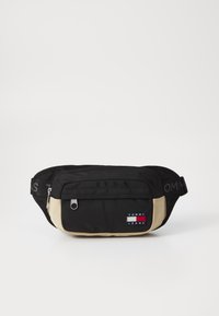 Fanny pack en tissu noir et beige avec une poche frontale zippée, un patch logo, une bandoulière réglable et une texture lisse.