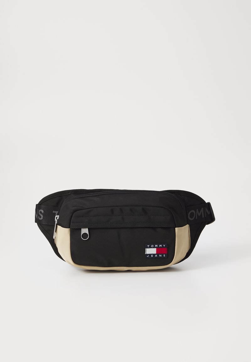 Fanny pack en tissu noir et beige avec une poche frontale zippée, un patch logo, une bandoulière réglable et une texture lisse.