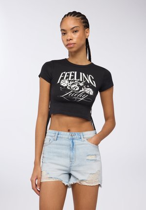 T-shirt nera cropped con testo e grafica stampati in bianco, abbinata a pantaloni shorts di jeans chiari con effetto usurato, orlo sfrangiato e tasche.