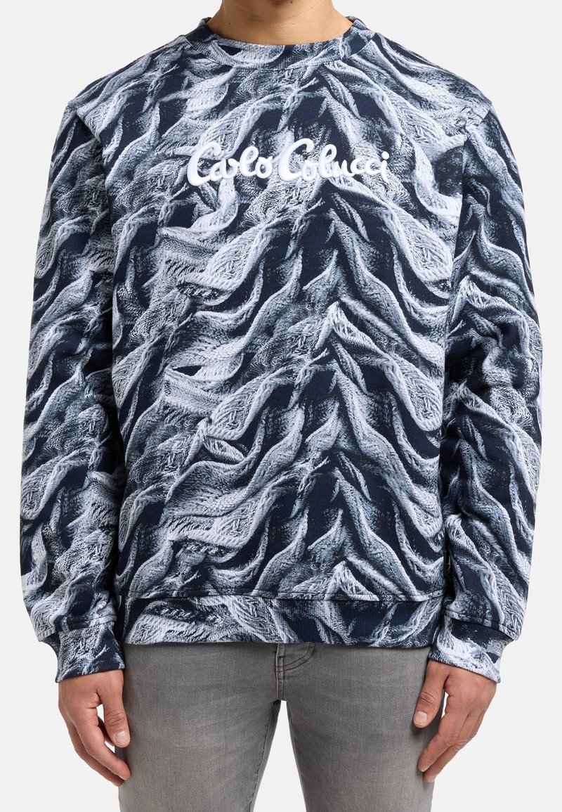 Homme portant un sweat-shirt texturé noir et blanc avec le logo « Carlo Colucci » en cursive blanche sur la poitrine, associé à un pantalon gris.