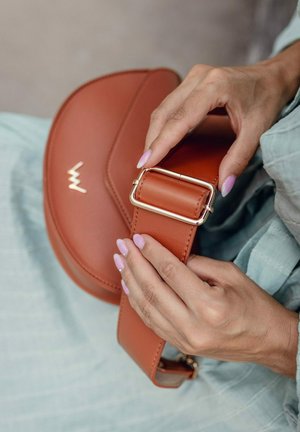 Hnědá kožená crossbody taška s zakřiveným tvarem. Obsahuje sponu z pozlaceného kovu a vystouplé logo. Hladká textura s elegantním designem.