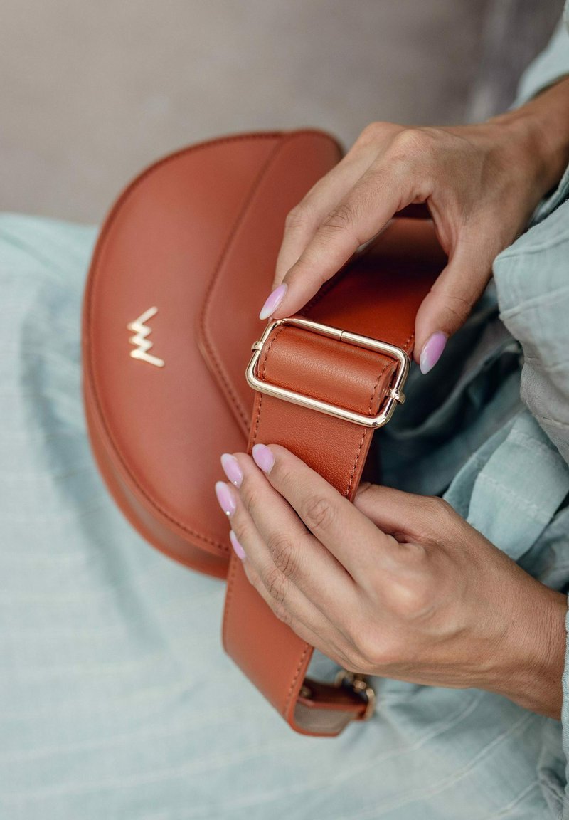 Hnědá kožená crossbody taška s zakřiveným tvarem. Obsahuje sponu z pozlaceného kovu a vystouplé logo. Hladká textura s elegantním designem.