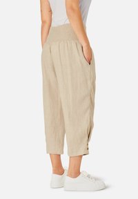 Žena nosící béžové lněné kalhoty do poloviny lýtek s nařaseným pasem na gumu a knoflíkovými detaily u lemu, v kombinaci s bílými teniskami.