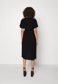 Black midi dress with short, flared sleeves, cinched waist, and a curved hem; paired with chunky black high-heeled sandals.