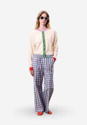 Femme portant un cardigan crème avec une bande de boutons verts, un col violet, des poignets rouges, un pantalon large à carreaux bleus, des chaussures rouges et des lunettes de soleil blanches.