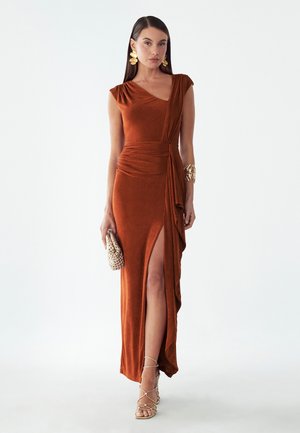 Femme portant une robe de soirée sans manches couleur rouille avec une fente haute, tenant une pochette perlée, accessoirisée avec des boucles d'oreilles et un bracelet en or.