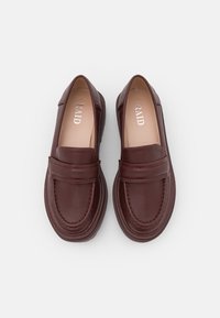Mocasines burdeos de cuero liso con costuras decorativas, una tira ancha y una suela baja y texturizada. Nombre de la marca "RAID" visible en el interior.