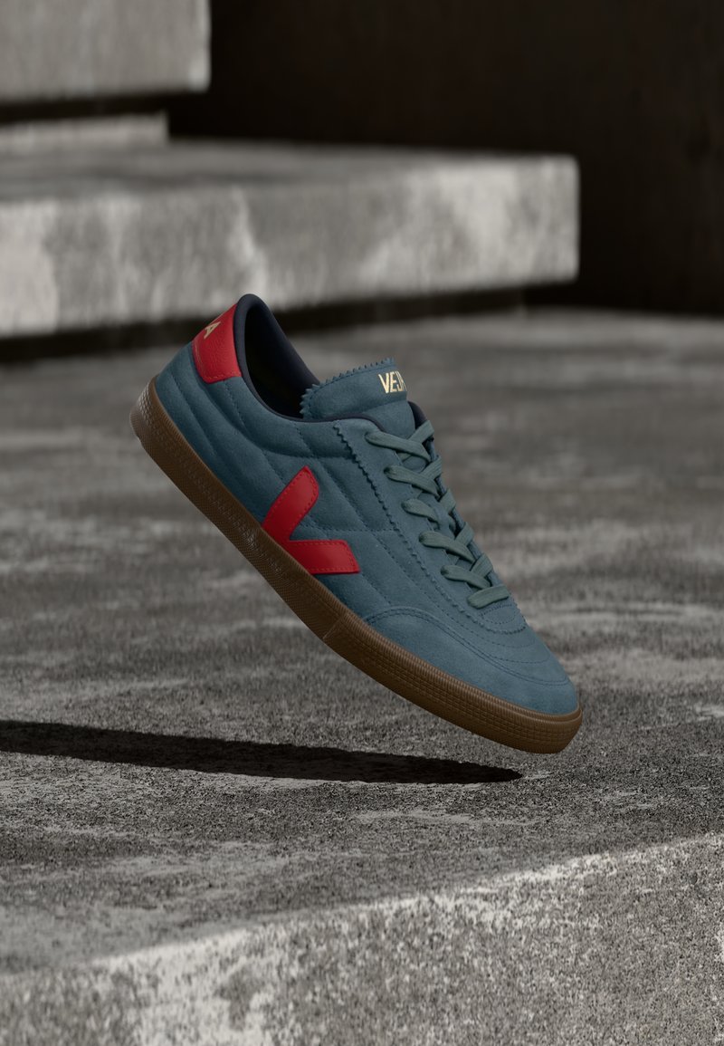 Μπλε αθλητικό παπούτσι με κόκκινες λεπτομέρειες και καφέ σόλα, αιωρούμενο πάνω από σκάλες από σκυρόδεμα με σκιά από κάτω.