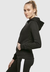 Hoodie noir en tissu doux, avec une poche frontale, une capuche à cordons et des poignets côtelés. Associé à un pantalon noir et blanc.